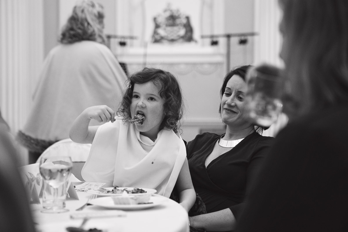 A black & white shot of a young girl scoffing cake while sat on her mothers lap at a wedding breakfast