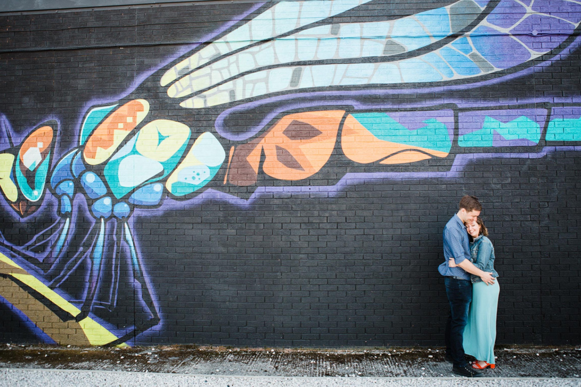 A young couple embrace white stood in front of a graffiti wall depicting a dragonfly
