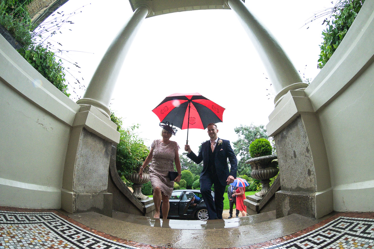 A wide, low angle shot of wedding guests arriving in the rain holding a black and red umbrella
