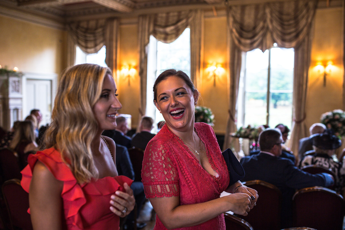 Two women wedding guests dressed in red laugh as they arrive at the ceremony.